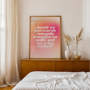 May include: A framed poster with a gradient background of pink and orange hues. The poster features the text: "Should we just search romantic comedies on Netflix and see what we find?" The poster is displayed above a wooden cabinet, with two vases and decorative plants.