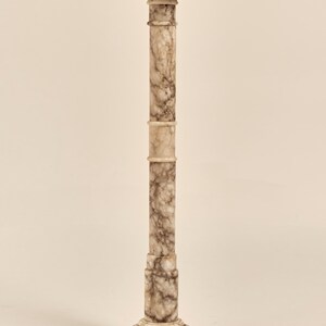 May include: A tall, marble pedestal with a square top and a decorative base. The column is a light beige color with dark brown veining throughout. The pedestal is a classic design, suitable for displaying sculptures or plants.