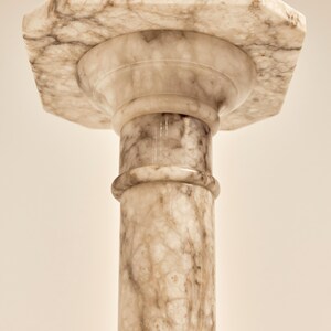 May include: A marble pedestal with a hexagonal top and a cylindrical column. The pedestal is a light beige color with darker brown veining throughout. The column has a rounded detail near the top.
