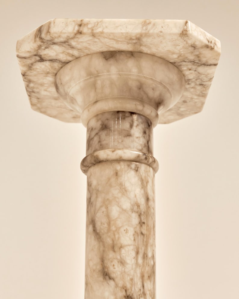May include: A marble pedestal with a hexagonal top and a cylindrical column. The pedestal is a light beige color with darker brown veining throughout. The column has a rounded detail near the top.