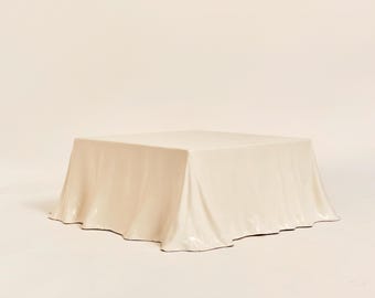 May include: A square, off-white coffee table with a draped, fabric-like appearance. The table has a smooth, glossy finish and a slightly wavy edge, creating a soft, elegant look. The table is set against a plain, light background.