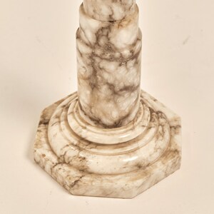 May include: A close-up of a marble pedestal with a hexagonal base. The pedestal has a light beige color with brown veining throughout. The design features a series of stacked cylindrical shapes.