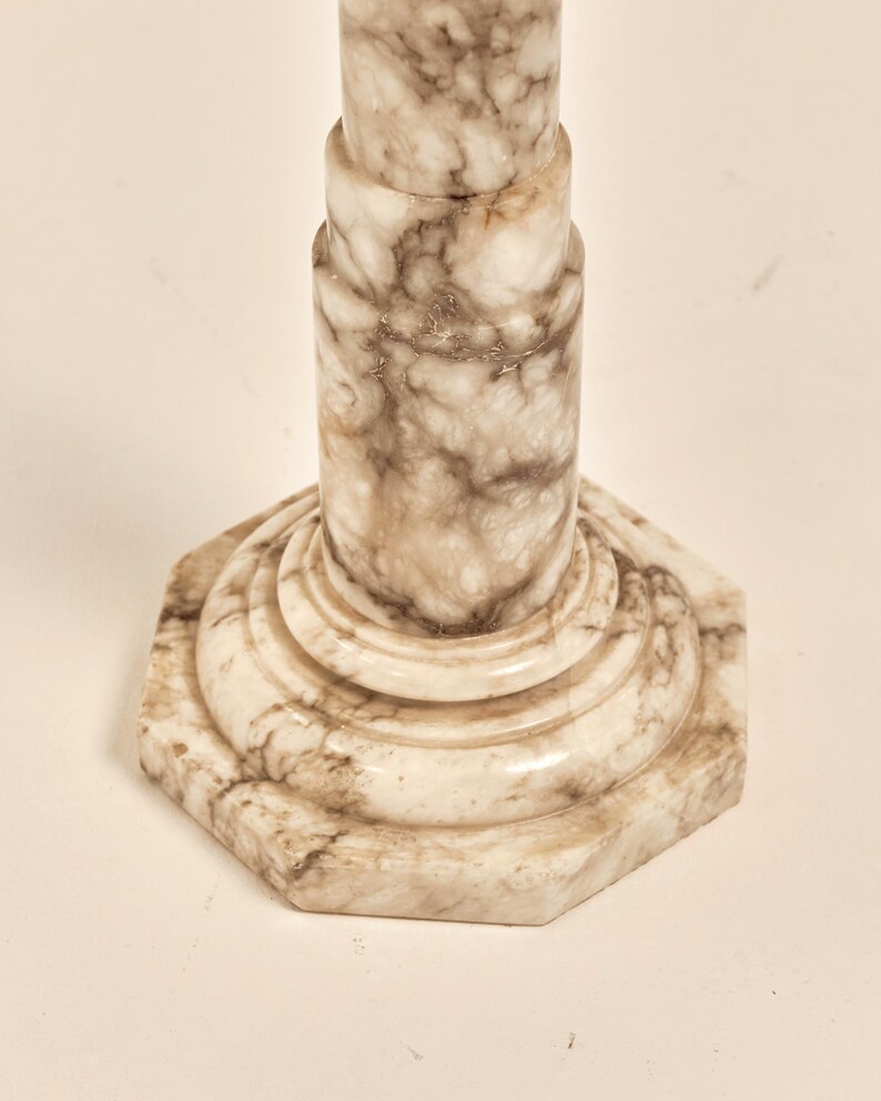 May include: A close-up of a marble pedestal with a hexagonal base. The pedestal has a light beige color with brown veining throughout. The design features a series of stacked cylindrical shapes.