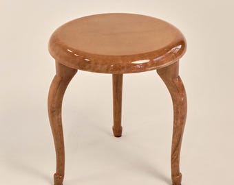 May include: A small, round stool with a glossy, light brown finish. The stool has a circular seat and three curved legs. The legs are also light brown and have a slight curve. The stool is a decorative furniture piece.