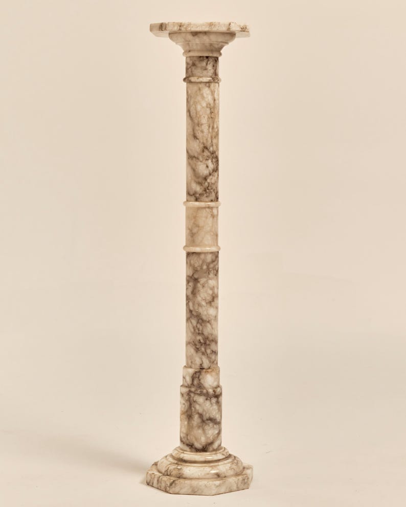 May include: A tall, marble pedestal with a square top and a decorative base. The column is a light beige color with dark brown veining throughout. The pedestal is a classic design, suitable for displaying sculptures or plants.