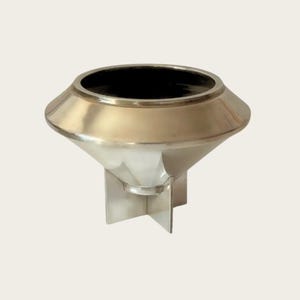 May include: A silver-colored metal decorative bowl with a wide, flared rim and a black interior. The bowl is supported by a cross-shaped base. The rim has a gold-colored band. The bowl has a modern, geometric design.