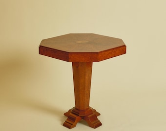 May include: An octagonal wooden side table featuring a rich brown burl wood veneer finish on the tabletop and central pedestal base. The table stands on a four-footed base with carved details, showcasing a smooth, polished surface throughout.