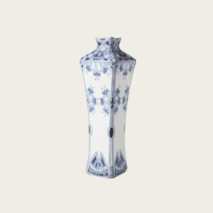 May include: A tall, white ceramic vase with a square base and a flared, geometric top. The vase is decorated with a blue floral pattern and vertical blue lines. The design is symmetrical and detailed, creating a classic, elegant look.
