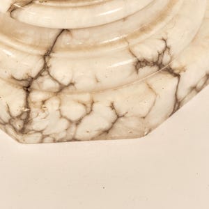 May include: Close-up of a decorative, off-white marble object with brown veining. The object has a rounded, tiered design with a hexagonal base. The marble has a polished, glossy finish.