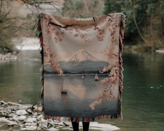 May include: A woven throw blanket depicting a landscape with a mountain, sailboats, and cherry blossoms. The blanket has a fringed edge with red, black, and tan accents. The image shows a tranquil outdoor scene.
