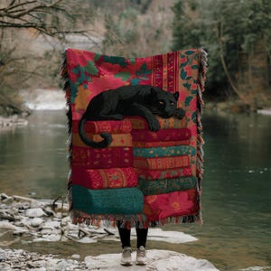 May include: A decorative throw blanket featuring a black panther resting on a stack of books. The blanket has a floral pattern in red, pink, and green. The fringed edges add a touch of elegance to this cozy home decor item.