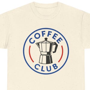 Könnte beinhalten: Cremefarbenes T-Shirt mit einem kreisförmigen Aufdruck. Der Aufdruck zeigt die Worte "COFFEE CLUB" und eine schwarz-weiße Kaffeemaschinen-Illustration, umrandet in Blau mit roten Akzenten. Das T-Shirt ist aus weichem Material.