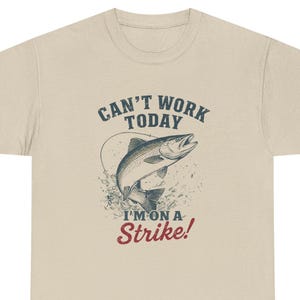 Könnte beinhalten: Beiges T-Shirt mit einer Grafik eines springenden Fisches und dem Text "CAN'T WORK TODAY I'M ON A Strike!". Das Design zeigt einen Fisch, der aus dem Wasser springt, mit einer Angelschnur. Das Shirt ist aus weichem, bequemem Material.