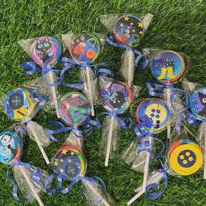 May include: A collection of round lollipops with colorful, whimsical designs, each individually wrapped and tied with a blue ribbon. The designs include a black cat, space scenes, and button graphics, set against a green grass background.