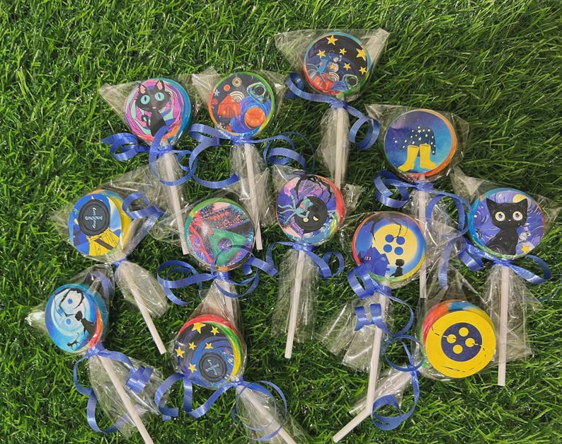 May include: A collection of round lollipops with colorful, whimsical designs, each individually wrapped and tied with a blue ribbon. The designs include a black cat, space scenes, and button graphics, set against a green grass background.