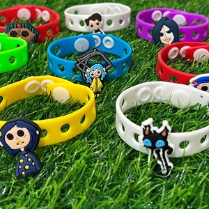 May include: A collection of colorful silicone bracelets with various character charms. The bracelets are in shades of red, pink, green, yellow, blue, purple, orange, white, and black. The charms feature cartoon characters. The bracelets are on a green surface.