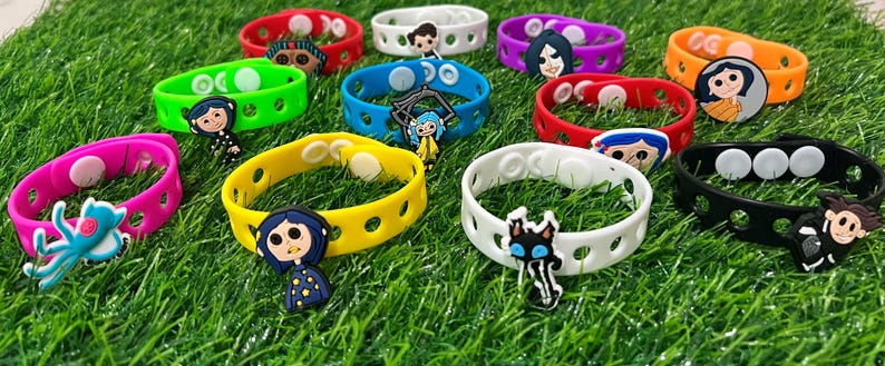 May include: A collection of colorful silicone bracelets with various character charms. The bracelets are in shades of red, pink, green, yellow, blue, purple, orange, white, and black. The charms feature cartoon characters. The bracelets are on a green surface.