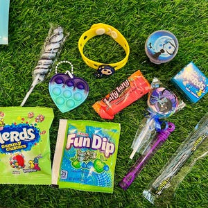 May include: A collection of colorful candies and toys on green grass. Items include a yellow bracelet, a heart-shaped pop-it, a swirl lollipop, a Fun Dip packet, and a Nerds gummy clusters bag.