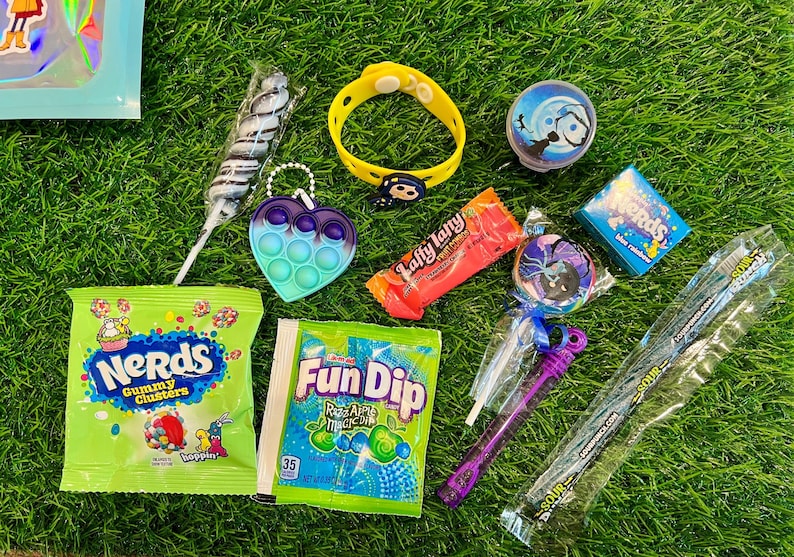 May include: A collection of colorful candies and toys on green grass. Items include a yellow bracelet, a heart-shaped pop-it, a swirl lollipop, a Fun Dip packet, and a Nerds gummy clusters bag.