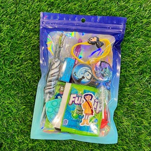 May include: A clear plastic bag filled with assorted colorful candies and small toys. Items include a swirl lollipop, a blue and green pop-it toy, and a packet of Fuzzi Razz Magic candy. A yellow bracelet with a black bow is also visible.
