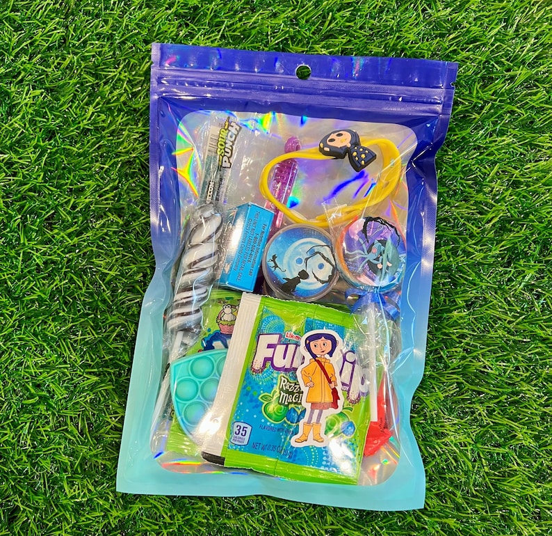 May include: A clear plastic bag filled with assorted colorful candies and small toys. Items include a swirl lollipop, a blue and green pop-it toy, and a packet of Fuzzi Razz Magic candy. A yellow bracelet with a black bow is also visible.