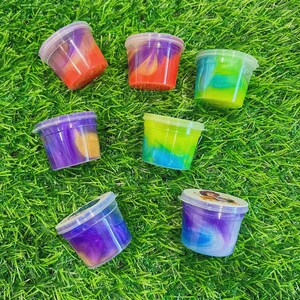 May include: Seven small plastic containers filled with colorful slime are arranged on a bed of green artificial grass. The slime colors include red, orange, purple, blue, and green, with some containers showing a swirl effect.