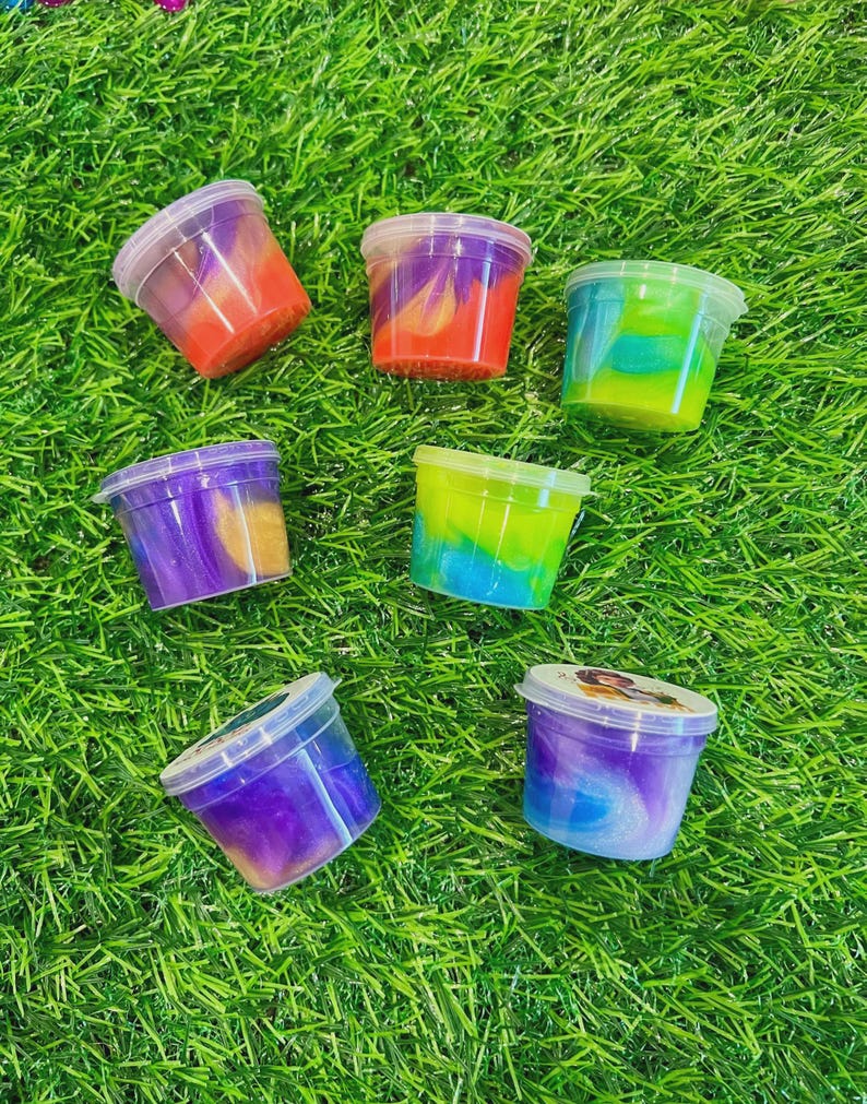 May include: Seven small plastic containers filled with colorful slime are arranged on a bed of green artificial grass. The slime colors include red, orange, purple, blue, and green, with some containers showing a swirl effect.