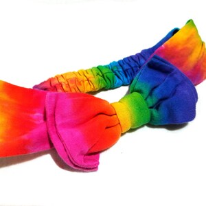 May include: A tie-dye headband in a vibrant rainbow of colors, including yellow, orange, pink, red, blue, and green. The headband features a knotted bow design and an elasticated band for a comfortable fit.