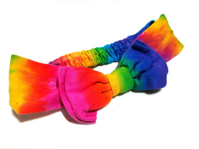 May include: A tie-dye headband in a vibrant rainbow of colors, including yellow, orange, pink, red, blue, and green. The headband features a knotted bow design and an elasticated band for a comfortable fit.