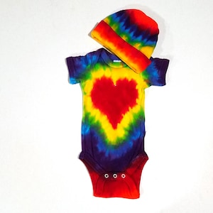 May include: A rainbow tie-dye baby onesie with a red heart design. The onesie is paired with a matching rainbow tie-dye beanie.