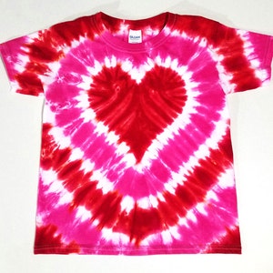 May include: A tie-dye t-shirt with a red heart design in the center. The shirt is white with pink and red dye swirling around the heart.