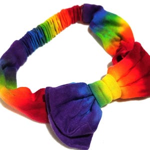 May include: A rainbow-colored headband with a knotted bow design. The headband transitions through the colors of the rainbow, from red to purple. The bow is a deep purple color.