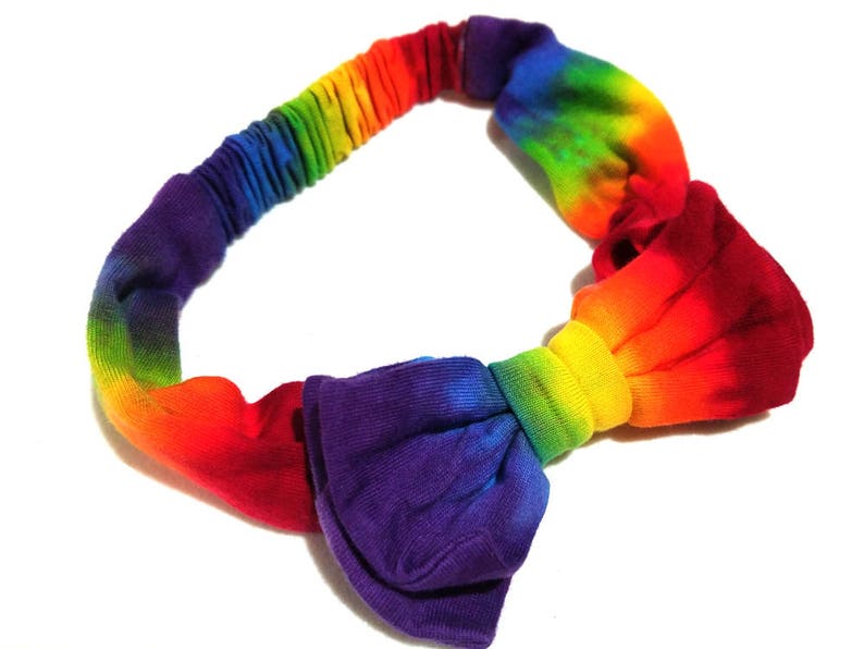 May include: A rainbow-colored headband with a knotted bow design. The headband transitions through the colors of the rainbow, from red to purple. The bow is a deep purple color.