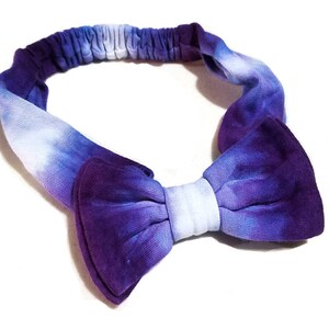 May include: A tie-dye headband in shades of purple and white. The headband features a bow in the center and an elastic band for a comfortable fit. The fabric has a soft, textured appearance.