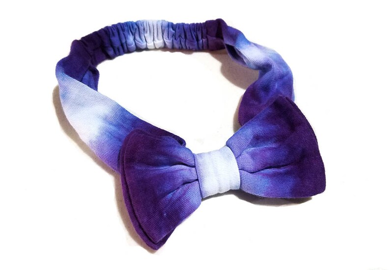 May include: A tie-dye headband in shades of purple and white. The headband features a bow in the center and an elastic band for a comfortable fit. The fabric has a soft, textured appearance.