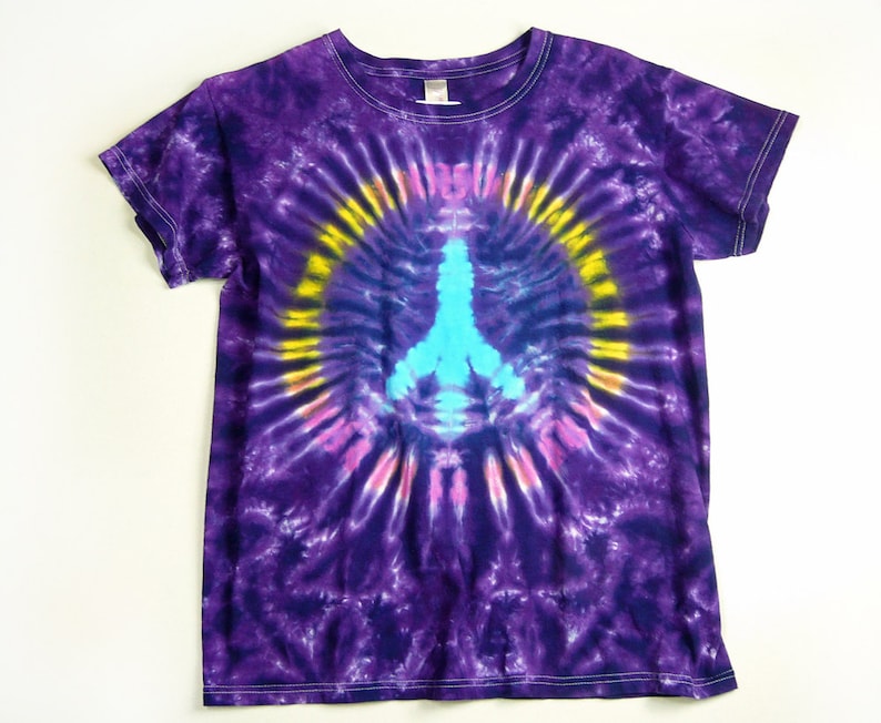Puede incluir: Camiseta de color morado con estampado tie-dye con un s&iacute;mbolo de paz azul en el centro. El s&iacute;mbolo de paz est&aacute; rodeado por un c&iacute;rculo amarillo y rosa.