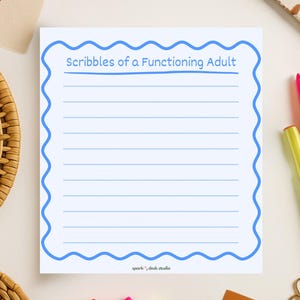 May include: A white notepad with a blue wavy border and the text "Scribbles of a Functioning Adult". The notepad has lined pages and is surrounded by colorful pens and office supplies.