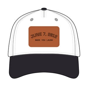 May include: A white baseball cap with a black brim. The cap features a brown rectangular patch with the text "JUNE 7, 2012 MADE YOU LAUGH" in black lettering. The cap has a black button on top.