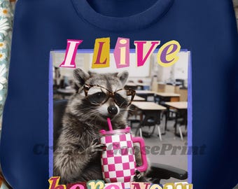 May include: Navy blue sweatshirt featuring a raccoon wearing glasses and drinking from a pink and white checkered cup. The text "I Live here Now" is displayed in colorful letters. The design is set against a classroom background.