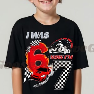 May include: Black t-shirt with a racing theme. The shirt features the text "I WAS 6 NOW I'M 7" with a large number 6 in red flames, a race car, and a checkered flag design. A speedometer and a race car are also visible.
