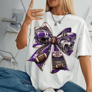 Peut inclure: Un t-shirt blanc arborant un grand motif de nœud violet à imprimé léopard. Le design du nœud intègre un casque de football américain, une tête de loup grognant, deux ballons de football marron et un terrain avec des poteaux de but.