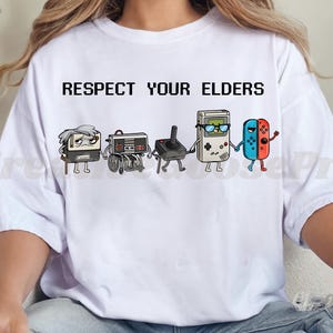 May include: White t-shirt with the text "RESPECT YOUR ELDERS" above a cartoon graphic of retro gaming consoles. The graphic includes a TV, a console on wheels, a joystick, a Game Boy, and a pair of Switch controllers.
