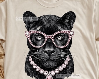 May include: A cream-colored t-shirt featuring a graphic print of a black panther wearing pink jeweled cat-eye glasses and a matching pink gemstone necklace. The panther has detailed fur texture and the text CreativeGoosePrint is visible.