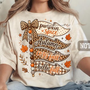 May include: A cream-coloured t-shirt featuring a graphic of four stacked pennant flags with the text pumpkin spice, Autumn, cozy fall, and hello fall. The design includes a vertical string of beads with a leopard print bow and orange maple leaves.