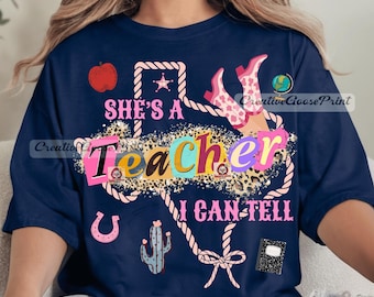 May include: A navy blue t-shirt featuring a graphic design with the text "SHE'S A TEACHER I CAN TELL". The design includes a rope border, a red apple, a globe, a cactus, a horseshoe, a notebook, and pink cowgirl boots with a leopard print pattern.