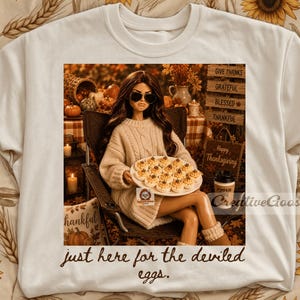 May include: A cream-coloured t-shirt featuring a graphic of a doll in a cable-knit sweater and sunglasses holding a plate of deviled eggs. The text reads: Give Thanks, Grateful, Blessed, Thankful, Happy Thanksgiving, and just here for the deviled eggs.