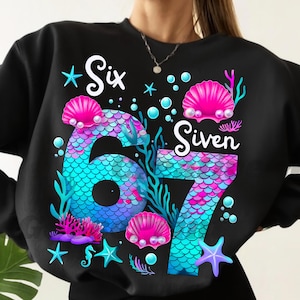 May include: Black sweatshirt with a colorful mermaid-themed design. The design features the numbers "6" and "7" in a mermaid scale pattern, surrounded by seashells, starfish, and the words "Six" and "Siven".