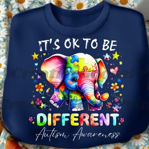 May include: Navy blue sweatshirt featuring a colorful elephant made of puzzle pieces. The text "IT'S OK TO BE DIFFERENT Autism Awareness" is displayed in a rainbow font, surrounded by stars and flowers.