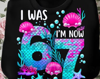 May include: Black sweatshirt with the text "I WAS 6, I'M NOW 7" in white. The numbers are designed with a mermaid scale pattern in shades of blue and pink, surrounded by seashells, coral, and starfish.
