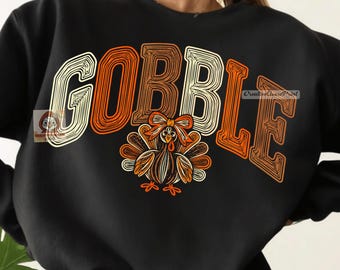 May include: A black long-sleeve sweatshirt featuring the word GOBBLE in large, stylized, multi-colored block letters. Below the text is a graphic of a turkey with orange, brown, and cream-colored feathers and a bow on its head.